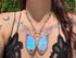 A close-up lifestyle shot of a person wearing a butterfly-shaped pendant necklace. The pendant features iridescent blue and green butterfly wings set in silver-toned metal and opal gemstone, suspended from a silver link chain.