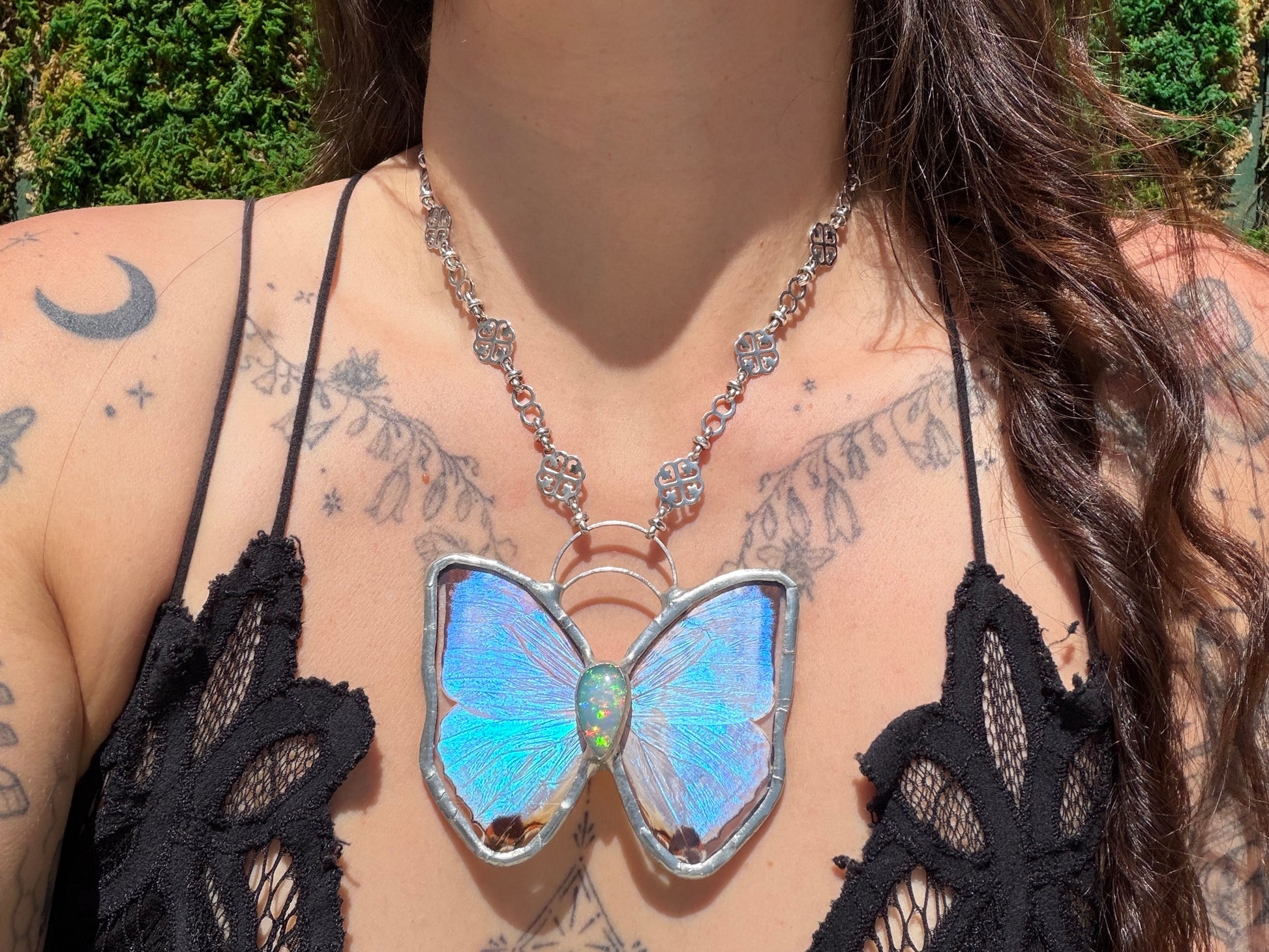 A close-up lifestyle shot of a person wearing a butterfly-shaped pendant necklace. The pendant features iridescent blue and green butterfly wings set in silver-toned metal and opal gemstone, suspended from a silver link chain.