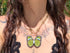 A close-up lifestyle shot of a person wearing a brass chain necklace with a butterfly pendant. The pendant features light green wings with blue and white accents, set against a background of the person's chest and hair.