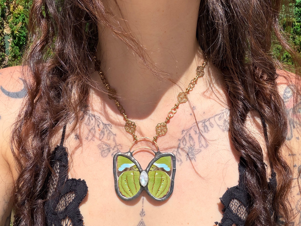 A close-up lifestyle shot of a person wearing a brass chain necklace with a butterfly pendant. The pendant features light green wings with blue and white accents, set against a background of the person's chest and hair.