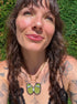 A close-up lifestyle shot of a woman with tattoos wearing a brass chain necklace with a butterfly-shaped pendant. The pendant features green and light blue enamel wings with a moonstone.