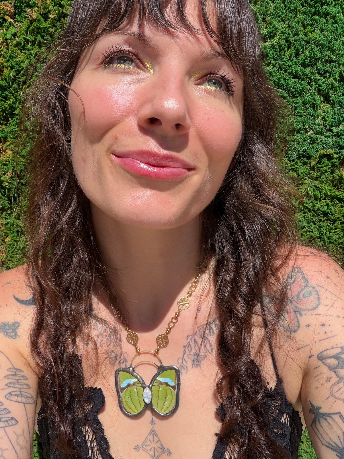 A close-up lifestyle shot of a woman with tattoos wearing a brass chain necklace with a butterfly-shaped pendant. The pendant features green and light blue enamel wings with a moonstone.