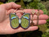 A hand holding a butterfly-shaped pendant necklace with green and blue inlay details and a brass chain and moonstone against a mossy green background.