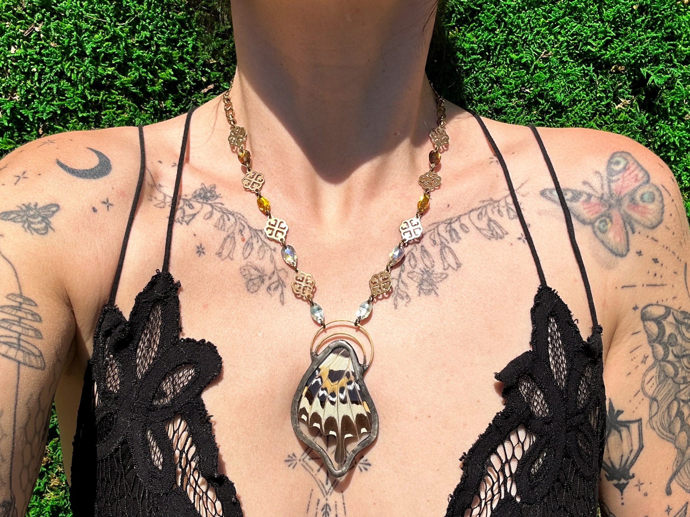 Person wearing a black lace top with a detailed butterfly necklace, standing against a green outdoor background.