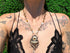 Person wearing a black lace top with a detailed butterfly necklace, standing against a green outdoor background.