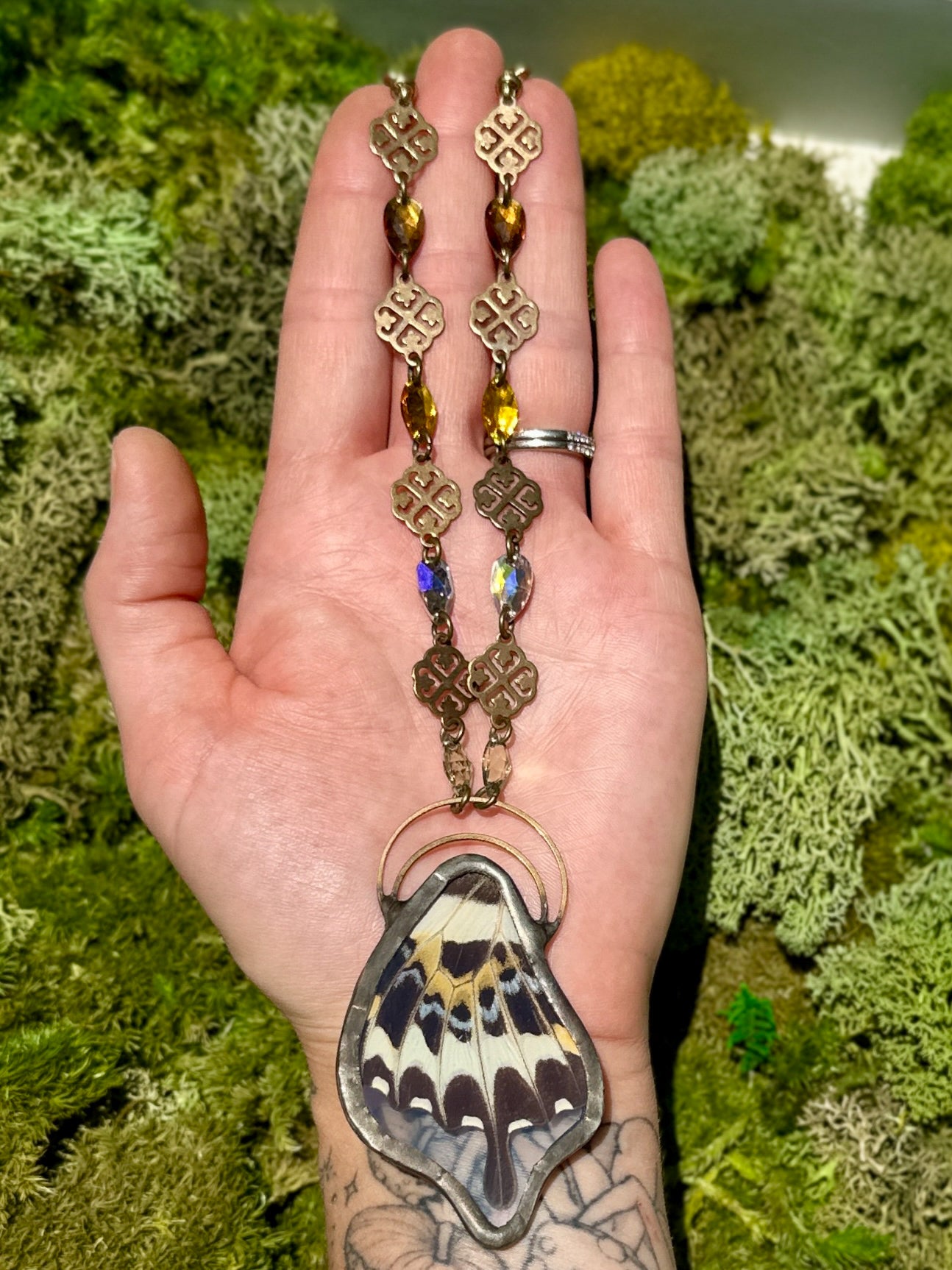 Hand holding a decorative necklace with a butterfly pendant against a green moss background