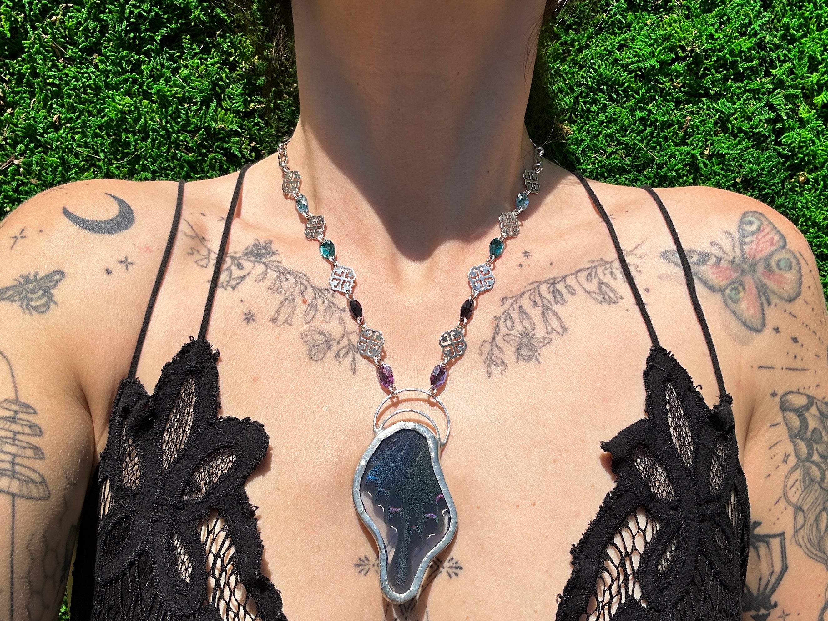 Person wearing a black lace top with a necklace featuring a large butterfly pendant, standing against a green outdoor background.