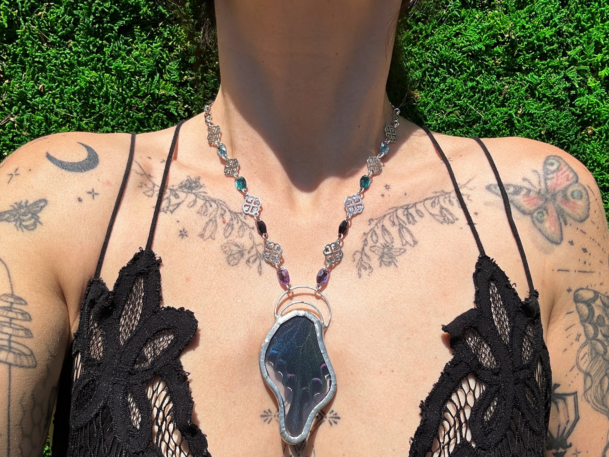 Person wearing a black lace top with a necklace featuring a large butterfly pendant, standing against a green outdoor background.