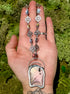 Hand holding a long, ornate necklace with a butterfly pendant against a green mossy background
