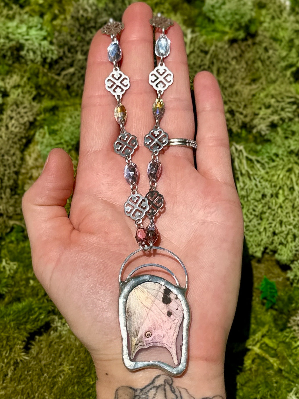 Hand holding a long, ornate necklace with a butterfly pendant against a green mossy background
