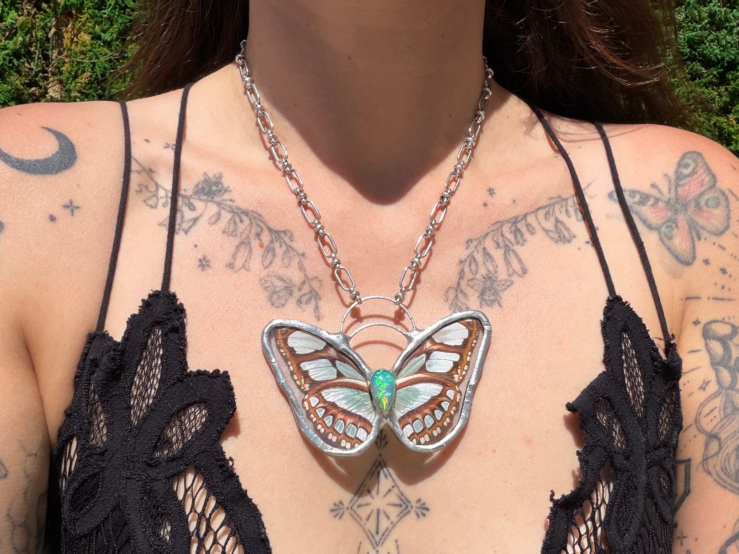 A close-up of a person wearing a silver butterfly pendant necklace with a opal gemstone, styled with a black lace top against an outdoor background.
