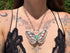 A close-up of a person wearing a silver butterfly pendant necklace with a opal gemstone, styled with a black lace top against an outdoor background.