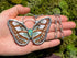A hand holds a butterfly silver pendant necklace with white and brown patterned wings and a iridescent opal gemstone, against a green mossy background.