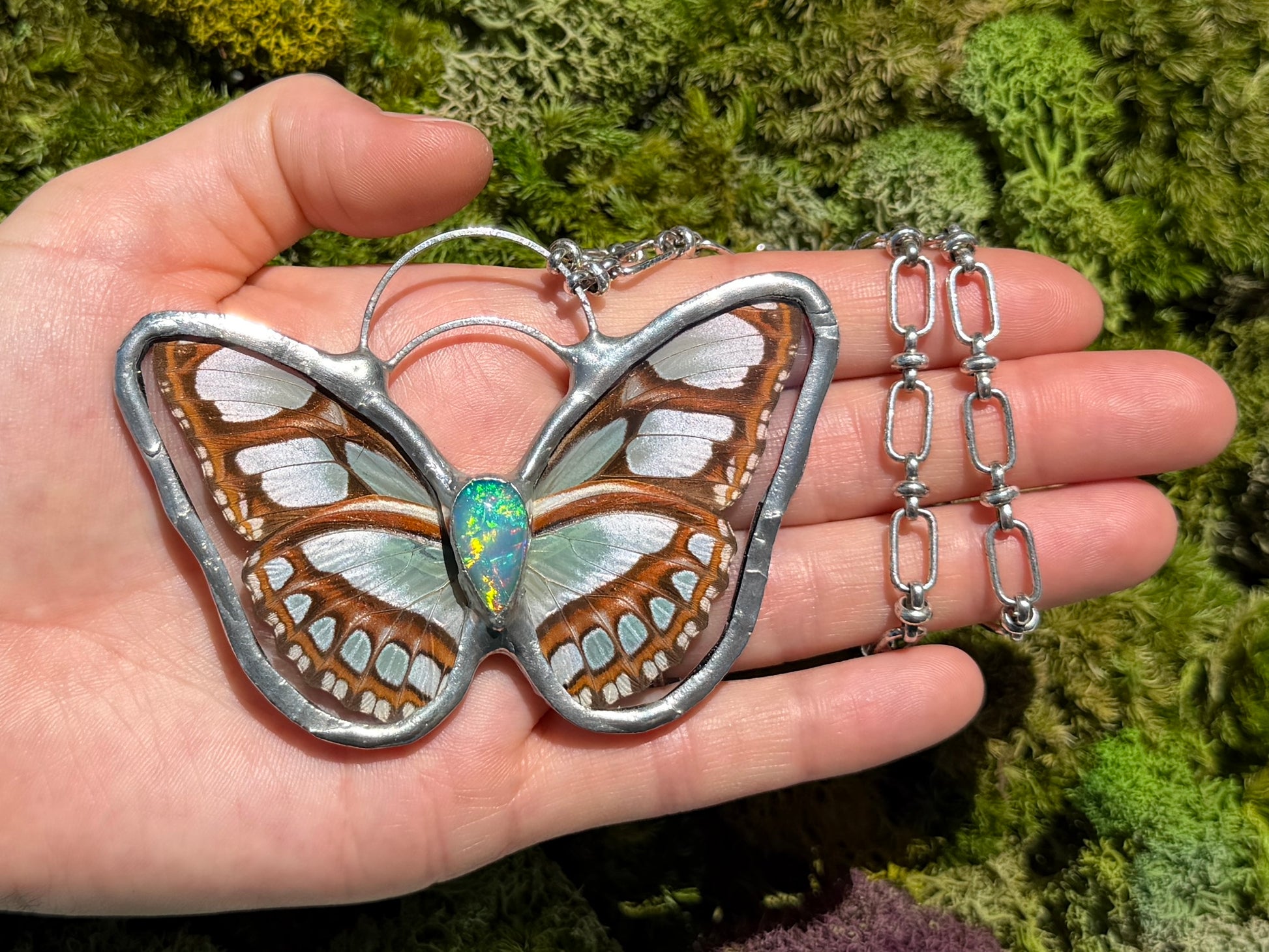 A hand holds a butterfly silver pendant necklace with white and brown patterned wings and a iridescent opal gemstone, against a green mossy background.
