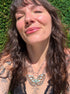 A close-up lifestyle shot of a woman with long dark wavy hair wearing a silver butterfly pendant necklace with turquoise and gold details, set against a green hedge background.