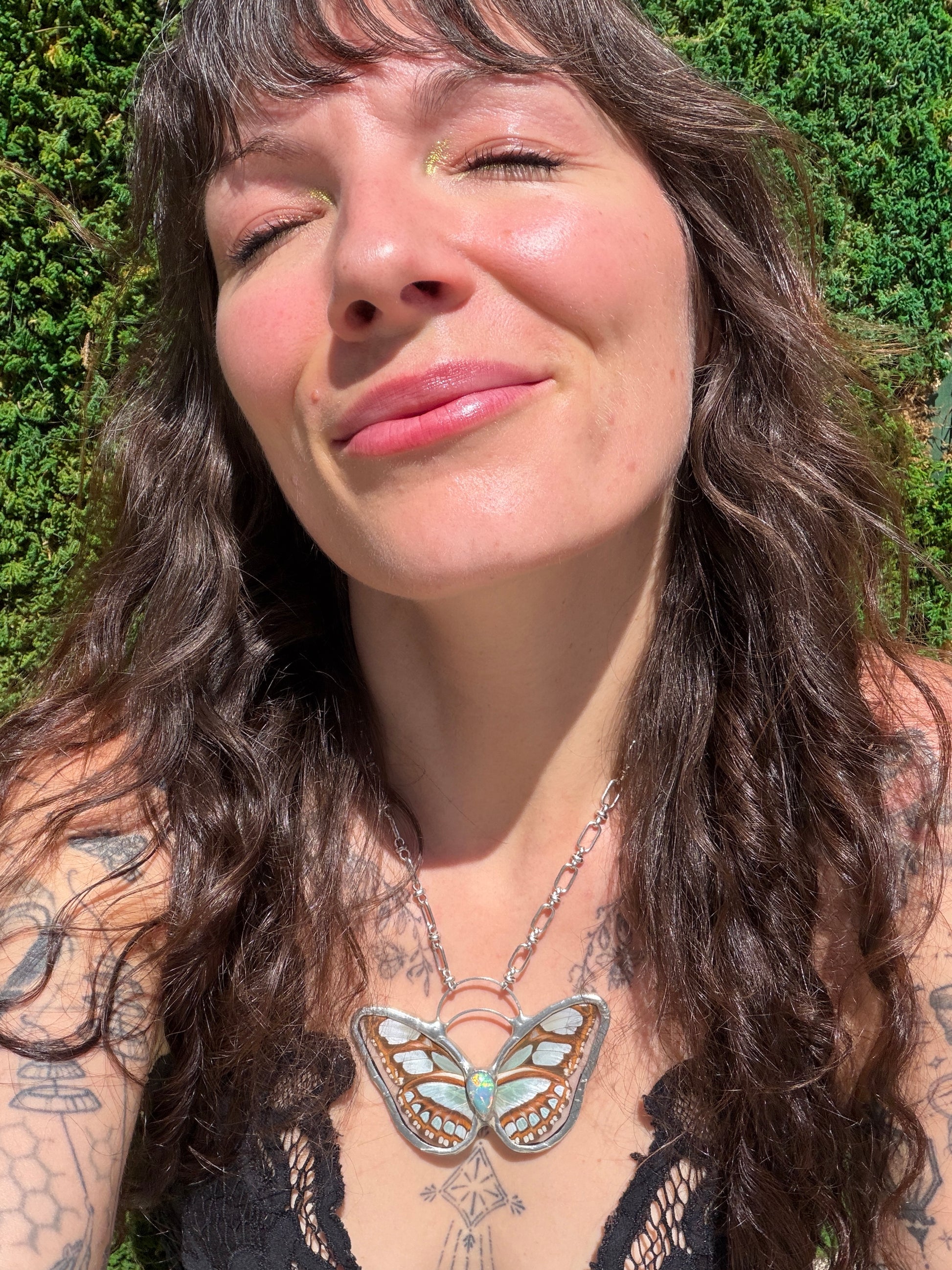 A close-up lifestyle shot of a woman with long dark wavy hair wearing a silver butterfly pendant necklace with turquoise and gold details, set against a green hedge background.