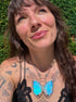 A close-up lifestyle shot of a woman wearing a silver butterfly pendant necklace with vibrant iridescent blue and green wings, with opal gemstone set against a background of green foliage.