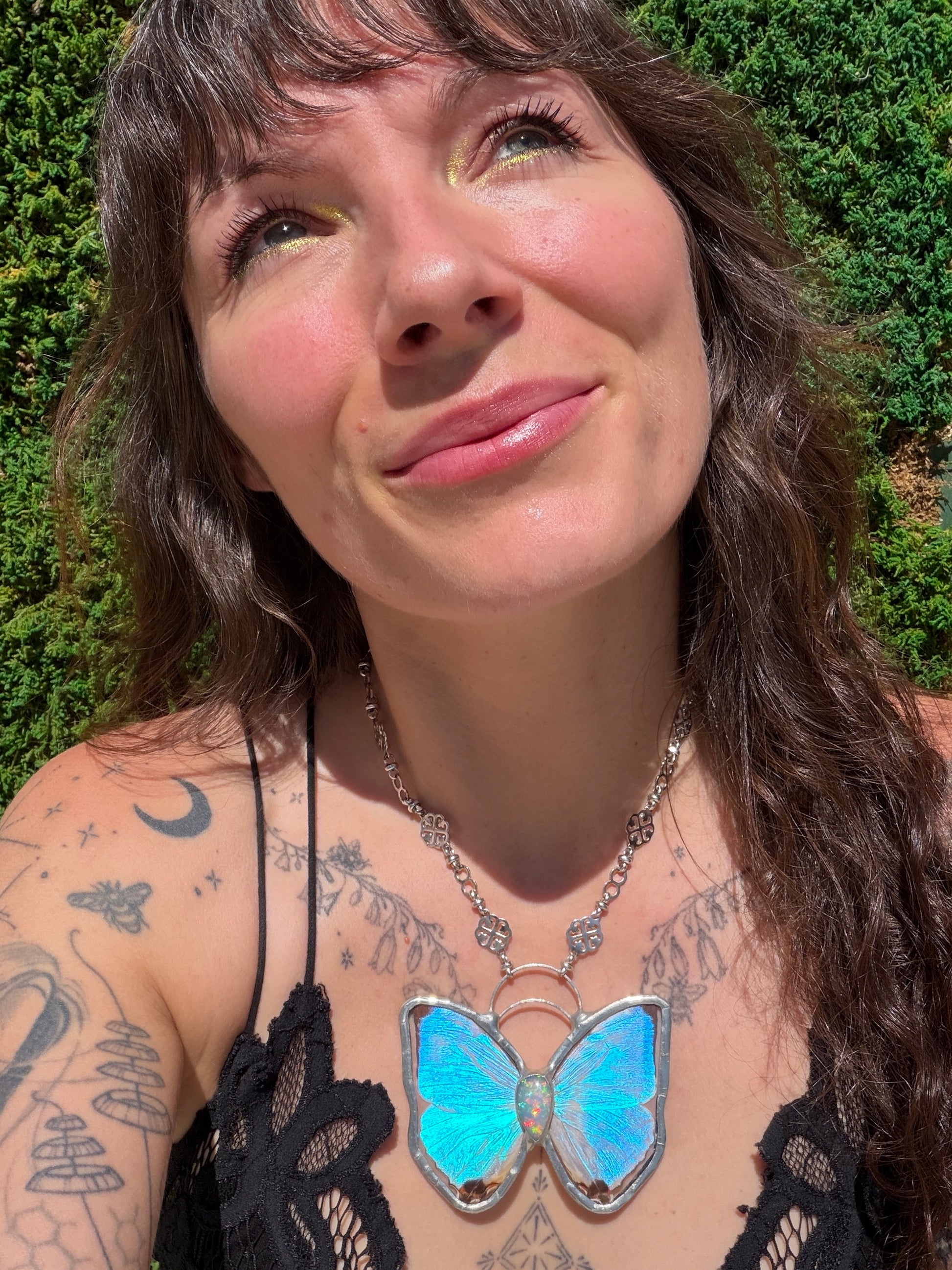 A close-up lifestyle shot of a woman wearing a silver butterfly pendant necklace with vibrant iridescent blue and green wings, with opal gemstone set against a background of green foliage.