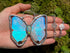 A hand holds a butterfly-shaped pendant necklace with iridescent blue and turquoise wings set in silver, alongside a matching silver chain and opal gemstone against a green mossy background.