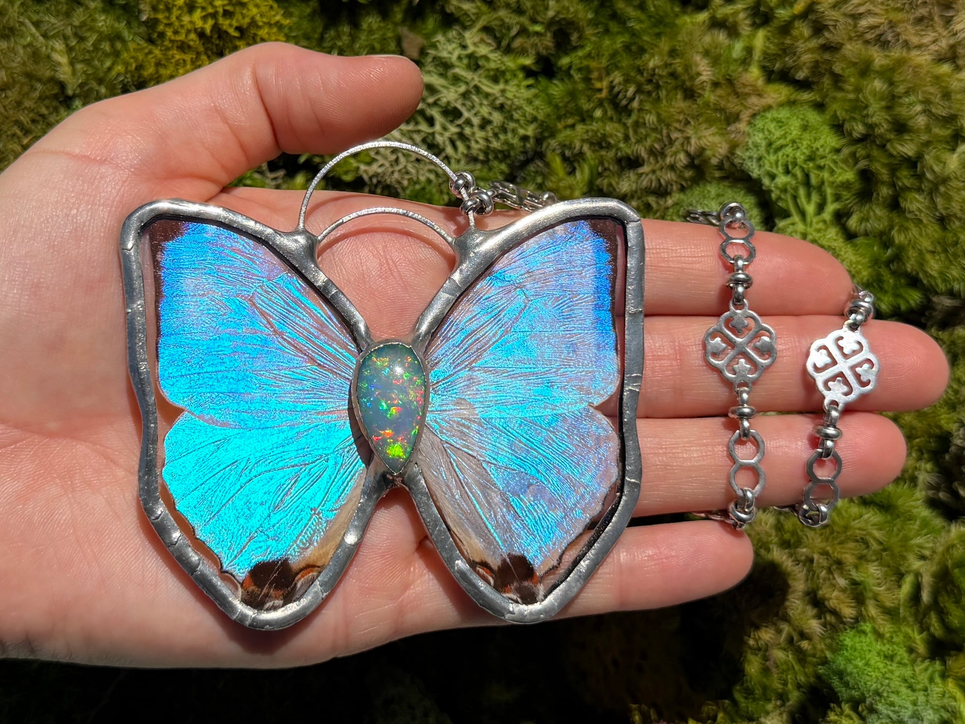 A hand holds a butterfly-shaped pendant necklace with iridescent blue and turquoise wings set in silver, alongside a matching silver chain and opal gemstone against a green mossy background.