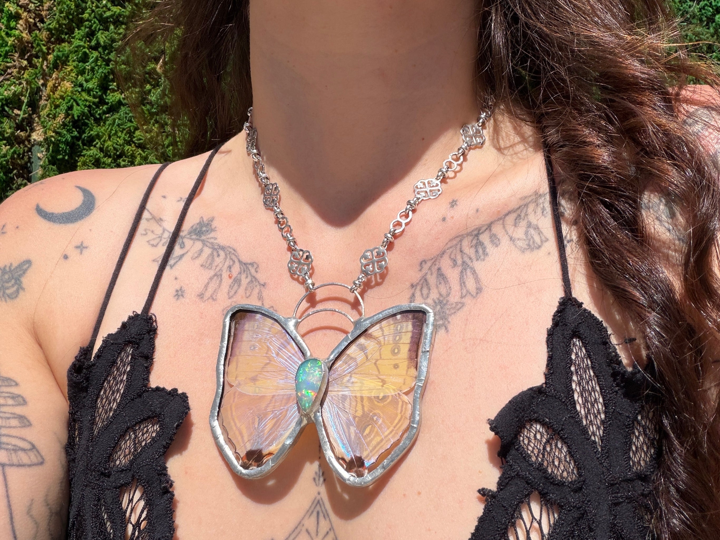 A close-up of a person wearing a silver butterfly pendant necklace with opal gemstone, iridescent yellow and blue wings, styled with a black lace top against a green outdoor background.