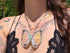 A close-up of a person wearing a silver butterfly pendant necklace with opal gemstone, iridescent yellow and blue wings, styled with a black lace top against a green outdoor background.
