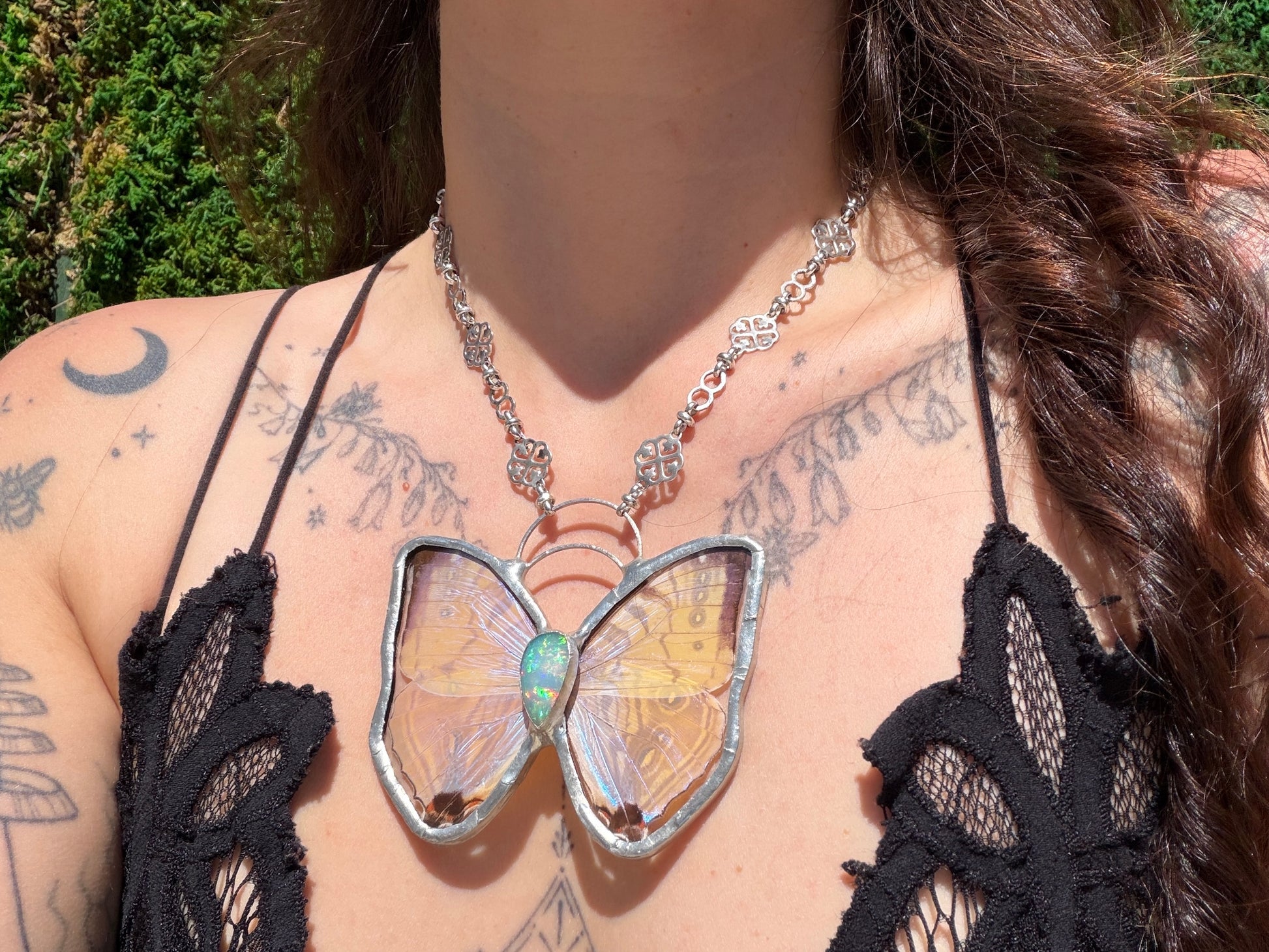A close-up of a person wearing a silver butterfly pendant necklace with opal gemstone, iridescent yellow and blue wings, styled with a black lace top against a green outdoor background.
