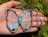 A hand holds a butterfly-shaped pendant necklace with iridescent wings and a silver-toned frame, radiant opal gemstone, alongside a matching silver chain, against a green mossy background.