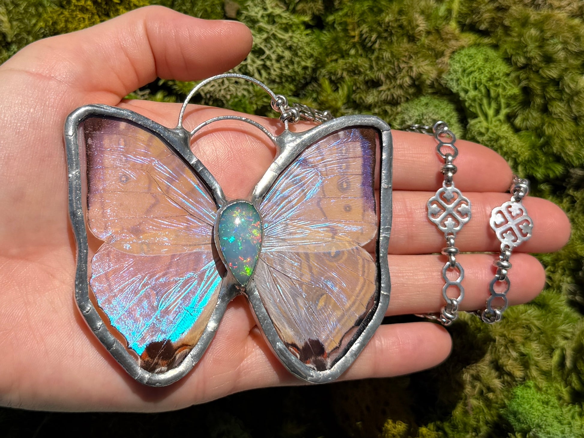 A hand holds a butterfly-shaped pendant necklace with iridescent wings and a silver-toned frame, radiant opal gemstone, alongside a matching silver chain, against a green mossy background.